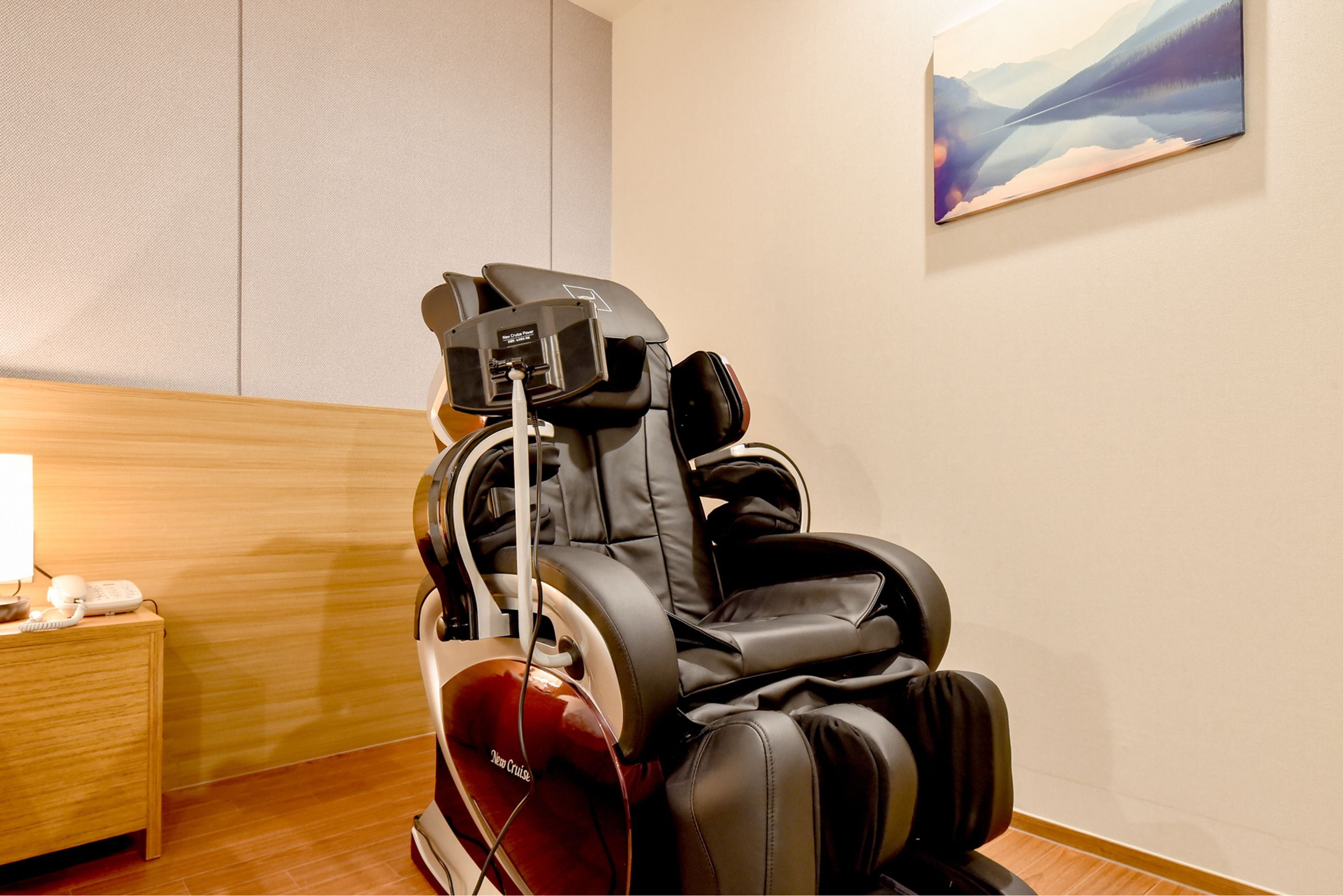 A massage chair in a room with a wooden floor, a small table with a lamp, and a painting on the wall.