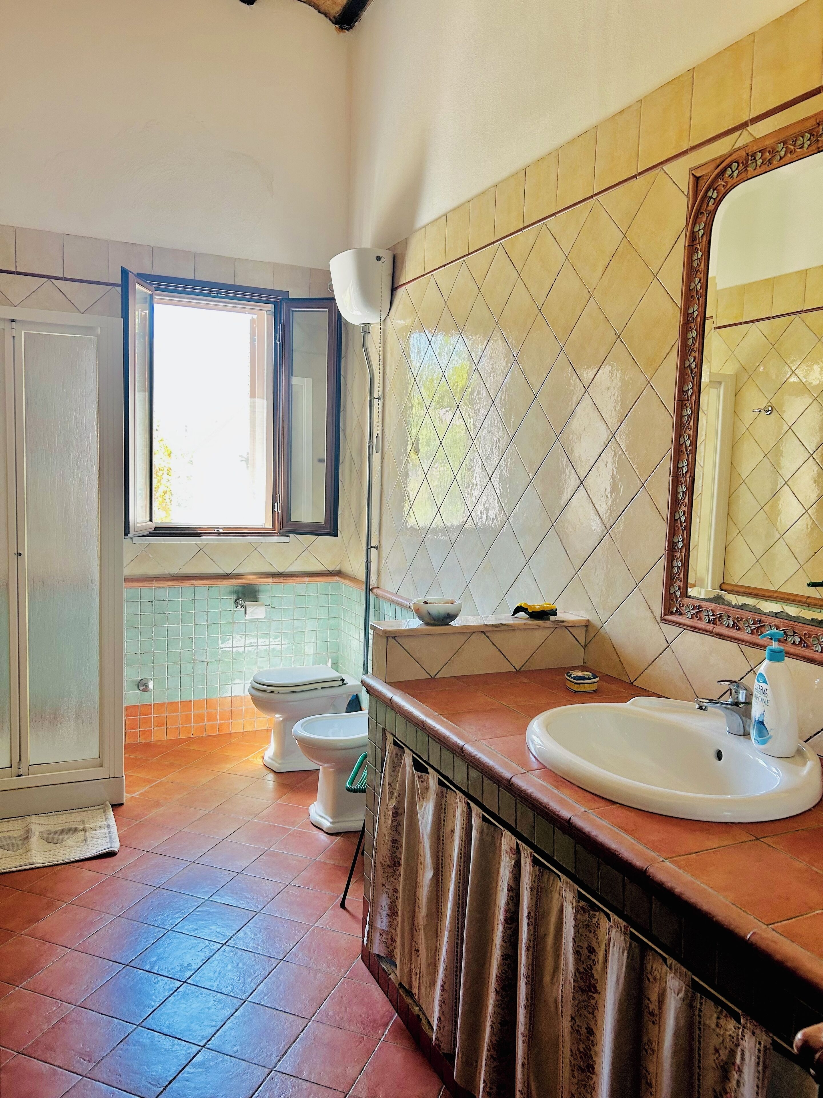 Un baño con inodoro, lavabo y espejo. Hay una ventana que proporciona luz natural.