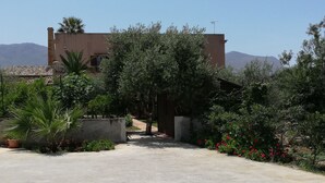 Un camino pavimentado que conduce a un edificio con tejado de azulejos, rodeado de vegetación y un telón de fondo montañoso.