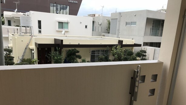 A balcony with a metal railing, a small trash bin, and a view of neighboring buildings. - Weekly Harbor View Mansion Ginowan (Ginowan)