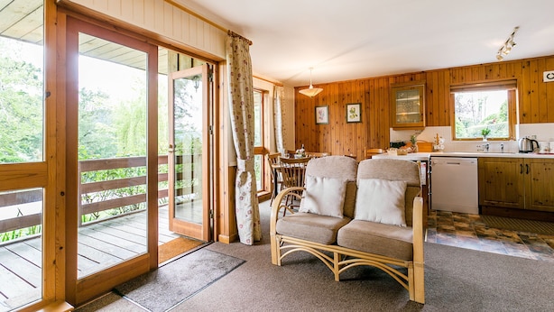 Et interiør af en træhytte med spiseplads, køkken og en balkon med udsigt over grøn bevoksning.