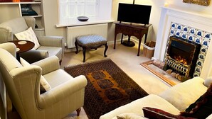 A living room with a sofa, armchair, and ottoman, a fireplace, a television, and a bookshelf with books and decorative items. - Grove Cottage -  a cottage that sleeps 4 guests  in 2 bedrooms (Yarmouth)