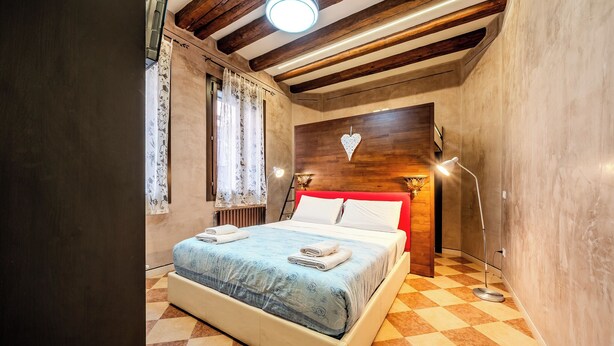 A bedroom with a checkered floor, a bed with a blue patterned bedspread, a wooden headboard with a heart decoration, and a standing floor lamp. - Campiello Tron Peaceful House (Venezia)