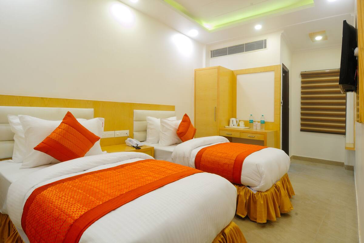 A hotel room with two beds, each with white bedding and orange accents, a wooden wardrobe, a mirror, and a television on the wall.