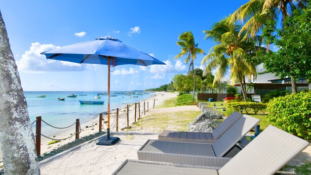 Eine Strandlounge mit Liegestühlen, einem blauen Sonnenschirm und Blick auf das Meer und Boote.