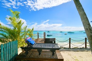 Una terraza de madera con una silla de descanso y una mesa, con vistas a una playa con barcos y palmeras.