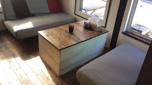 A cozy room with a wooden coffee table, a grey sofa with a red cushion, and a window with a view of the outside.