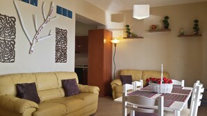 Una sala de estar con un sofá amarillo, una mesa de comedor con sillas blancas, decoración en la pared y una cocina al fondo.
