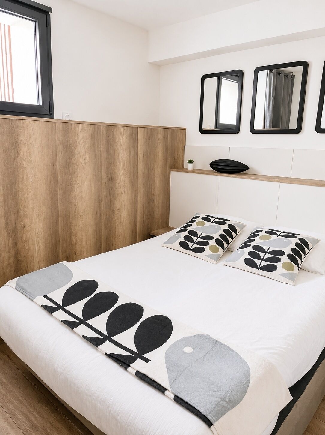 Ein Schlafzimmer mit einem Holzkopfteil, einem Bett mit gemusterten Kissen, einem Nachttisch mit einer Pflanze und drei Wandspiegeln.