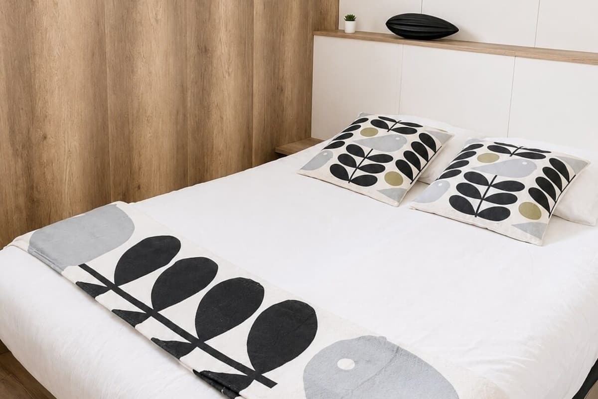 A bedroom with a wooden headboard, a bed with patterned pillows, a bedside table with a plant, and three wall mirrors.