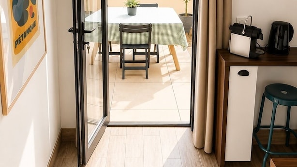 A room with a sliding glass door leading to an outdoor balcony with a table and chairs, a small kitchenette with a coffee maker, and a framed picture on the wall.