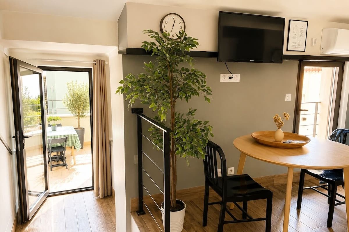 A modern living room with a dining table, a potted plant, a black chair, a wooden floor, a TV mounted on the wall, and a sliding glass door leading to an outdoor area.