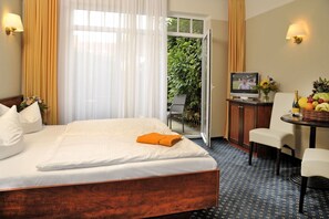 Habitación de hotel con una cama grande, un televisor sobre un mueble, una mesita con fruta y una botella, y vistas a zonas verdes a través de la puerta.