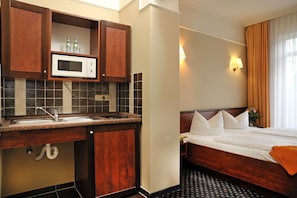 Quarto de hotel com cozinha compacta, cama com roupa de cama branca, mesa de cabeceira e janela com cortinas.