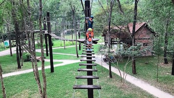 Una persona cruzando un puente colgante en una zona boscosa, con un edificio rústico al fondo.