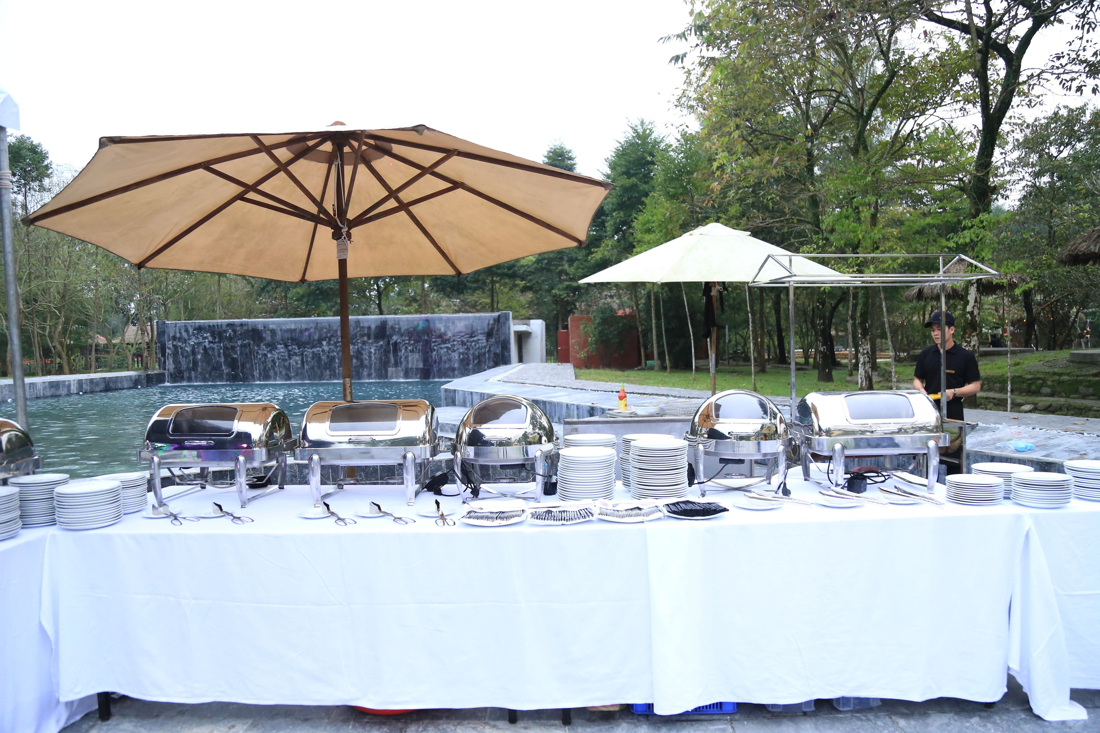 A buffet setup with multiple serving stations, a large umbrella, and a waterfall in the background.