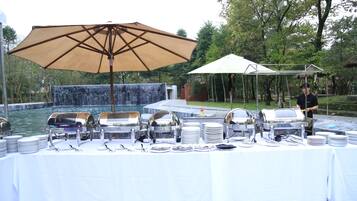 A buffet setup with multiple serving stations, a large umbrella, and a waterfall in the background.