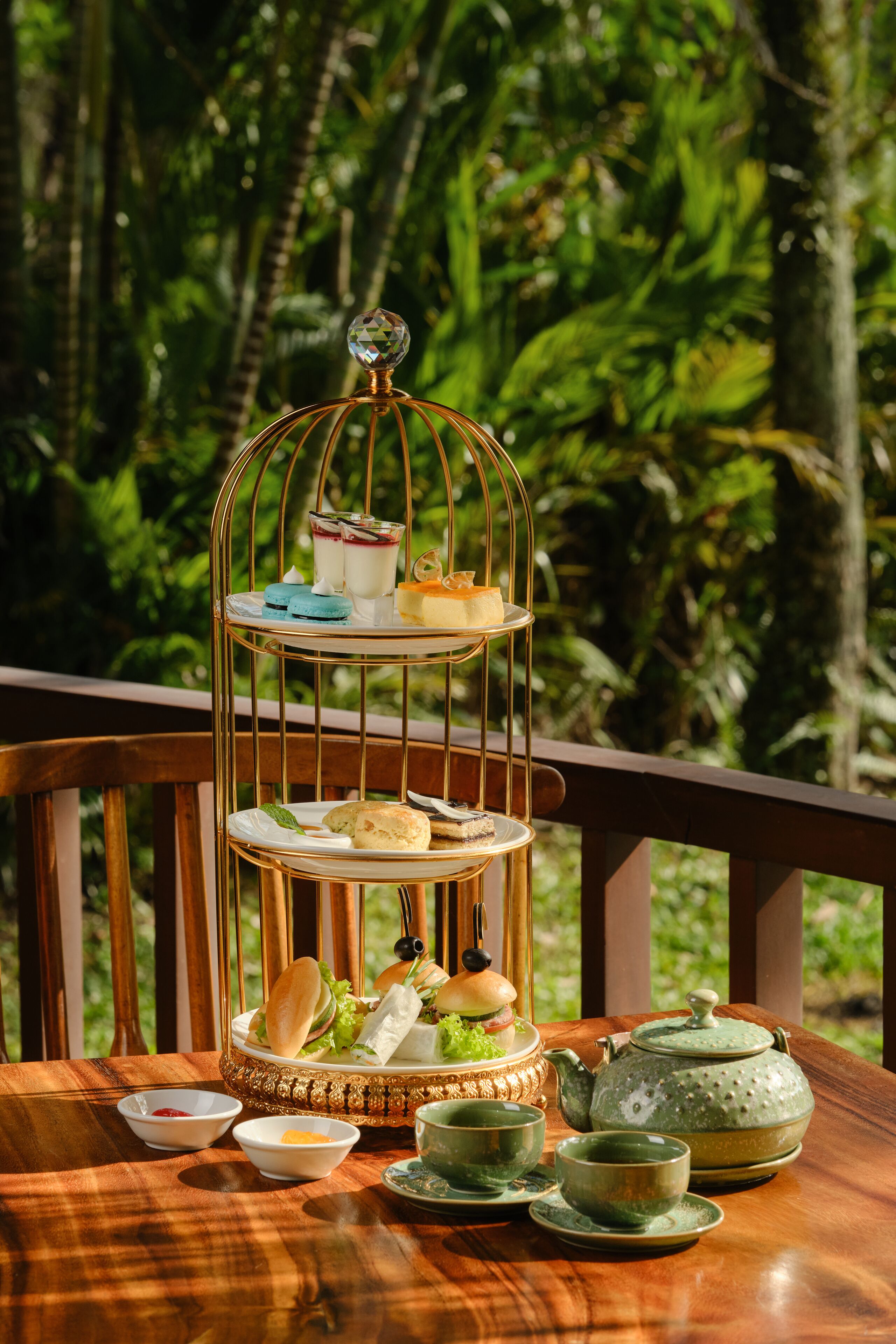 A tiered dessert stand with pastries and drinks on a wooden table, surrounded by a lush green forest.