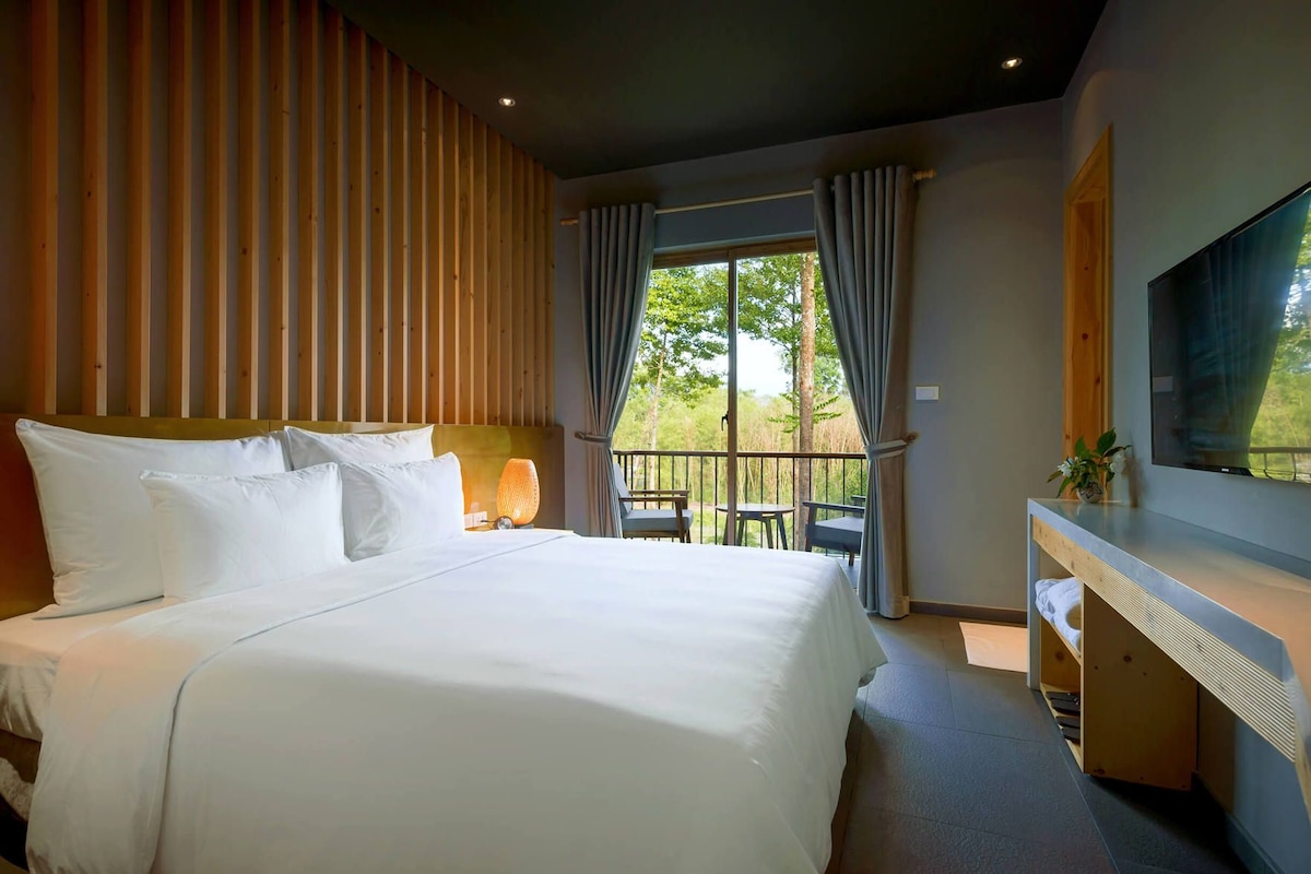 A hotel room with a bed, a desk, a TV, and a balcony with outdoor seating.