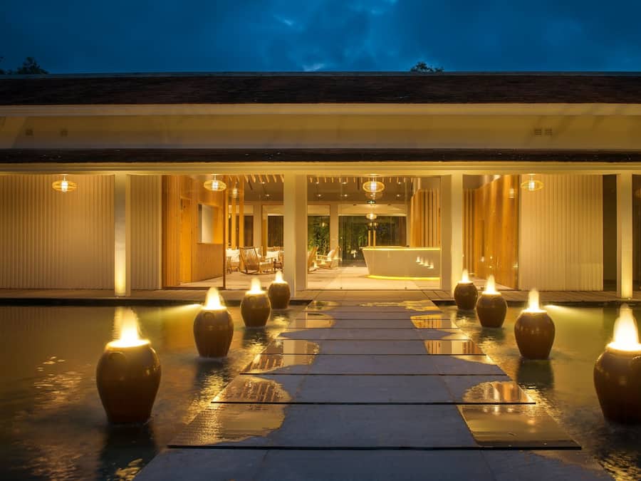 A modern hotel entrance with a reception desk, indoor seating, and a pathway lined with candles.