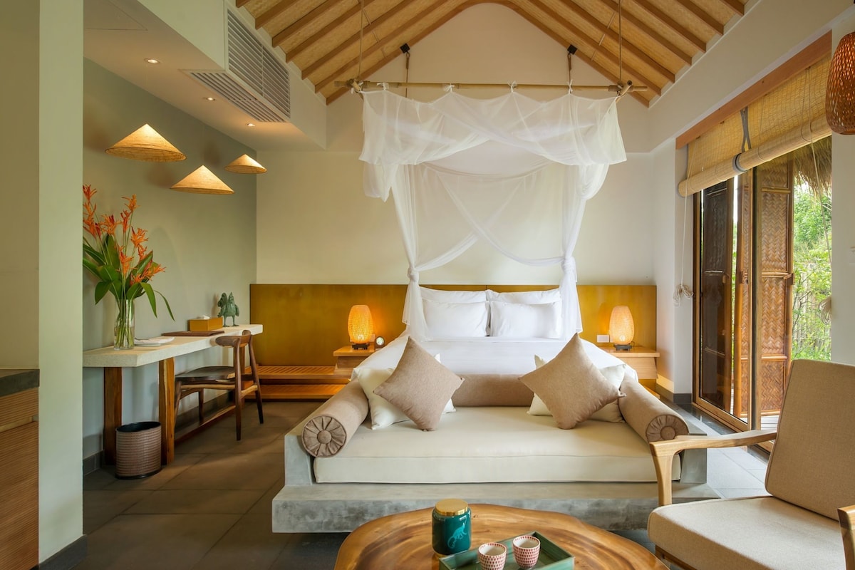 Une chambre moderne avec un lit à baldaquin, un coin salon avec un canapé et des fauteuils, une table à manger et une vue sur la verdure environnante.