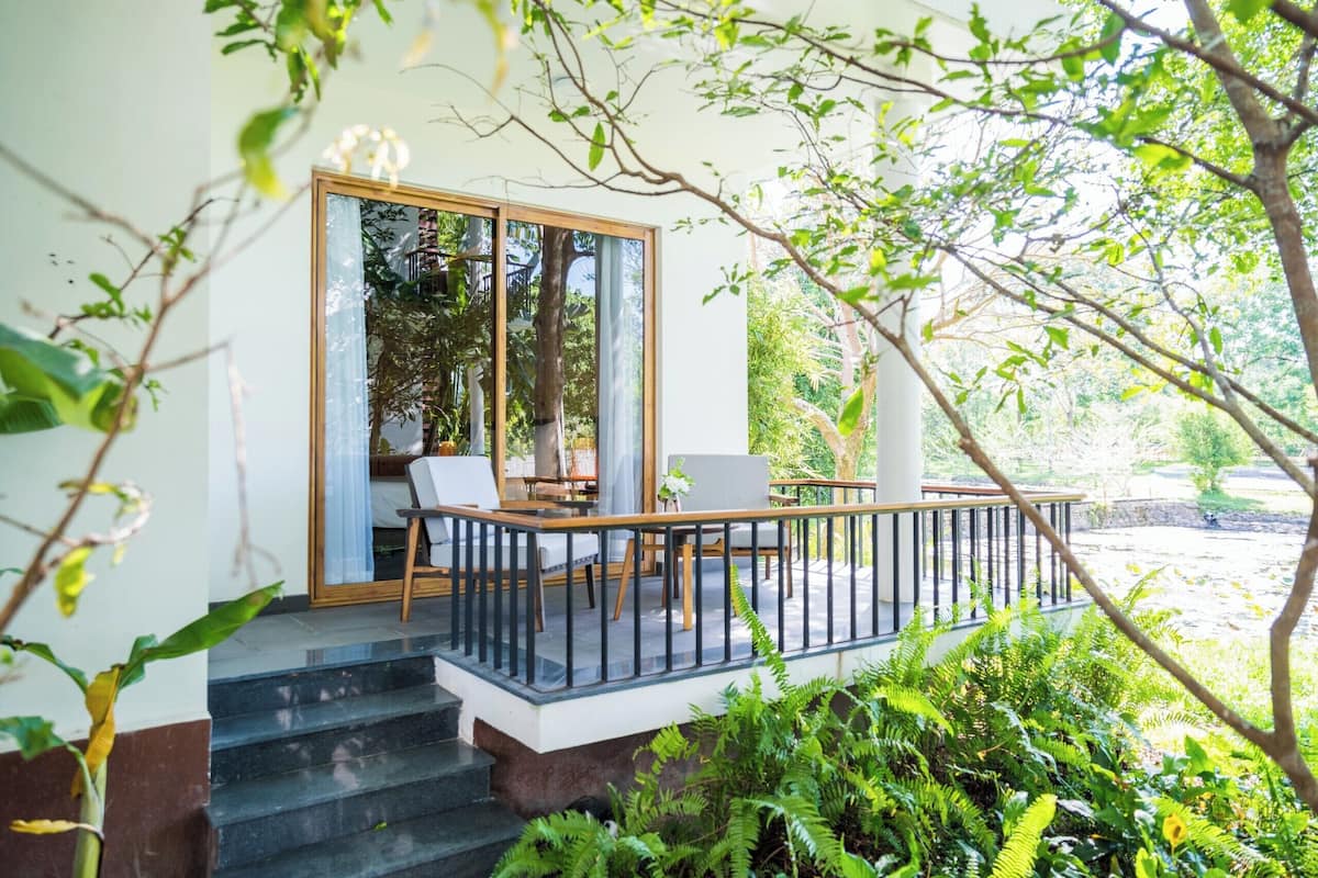 Un área de estar exterior moderna con barandilla de vidrio, cubierta de madera y abundante vegetación.
