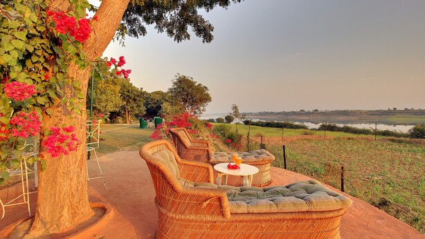 A wicker sofa set with cushions, a small round table, and a glass of orange juice on a patio with a view of a lake and fields. - Bhadrawati Safari Ranthambore Resort (Khandar)
