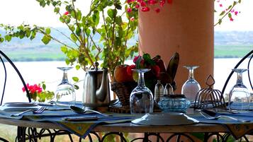 Tavola apparecchiata con diversi bicchieri, posate, un boccale di metallo e una pianta in vaso con foglie verdi e fiori rosa, posizionata all'aperto con vista su un corpo d'acqua sullo sfondo.