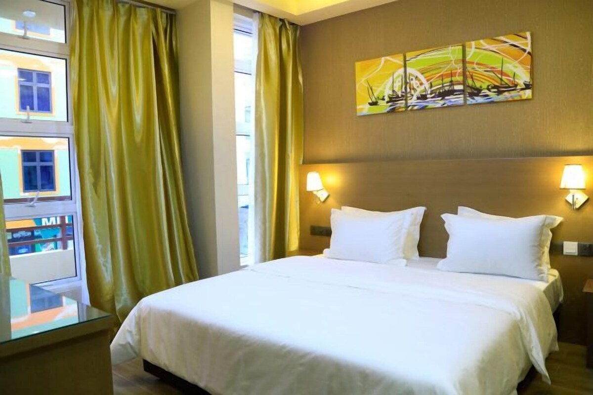 Quarto de hotel com uma cama grande, duas luminárias de cabeceira, um quadro na parede e vista para um edifício.