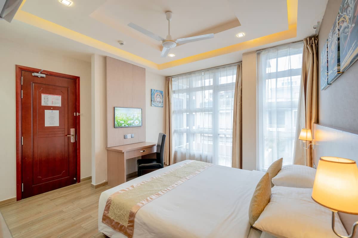 A hotel room with a bed, desk, chair, ceiling fan, and large windows with curtains.
