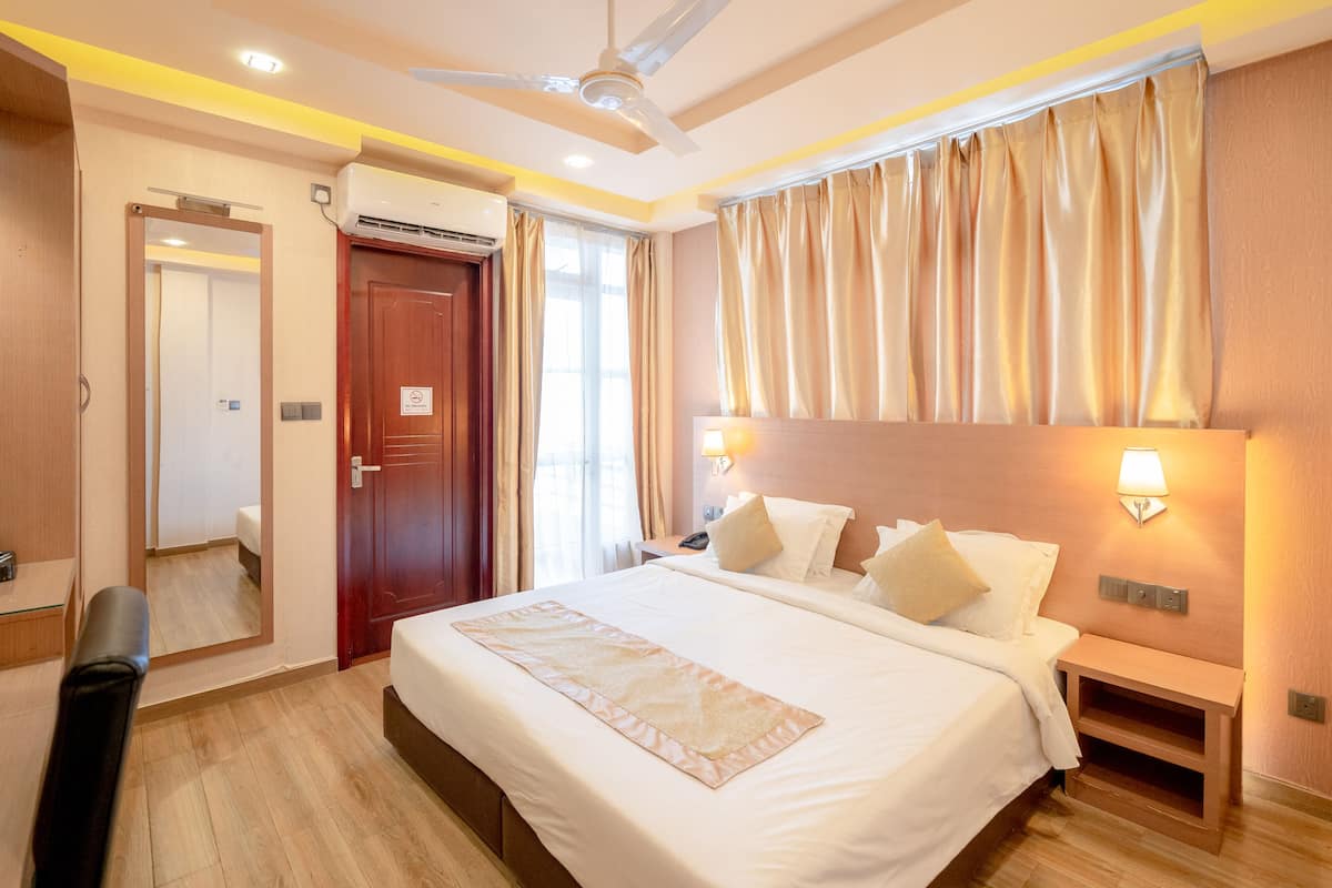A hotel room with a large bed, a ceiling fan, an air conditioning unit, a bedside table with a lamp, and a wooden floor.