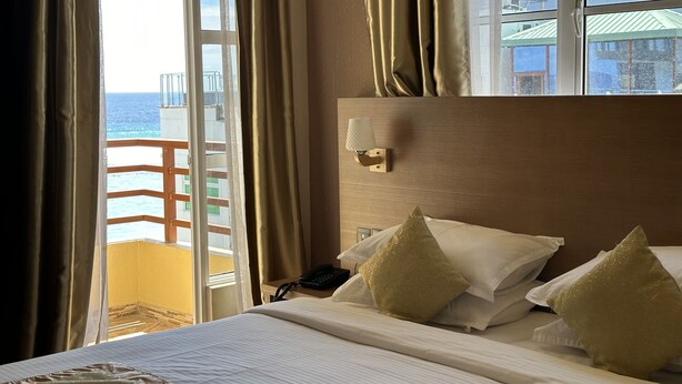 A hotel room with a bed, a nightstand, a lamp, and a view of the sea through a balcony door. - Three Inn (Hulhumalé)