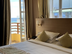 A hotel room with a bed, a nightstand, a lamp, and a view of the sea through a balcony door.