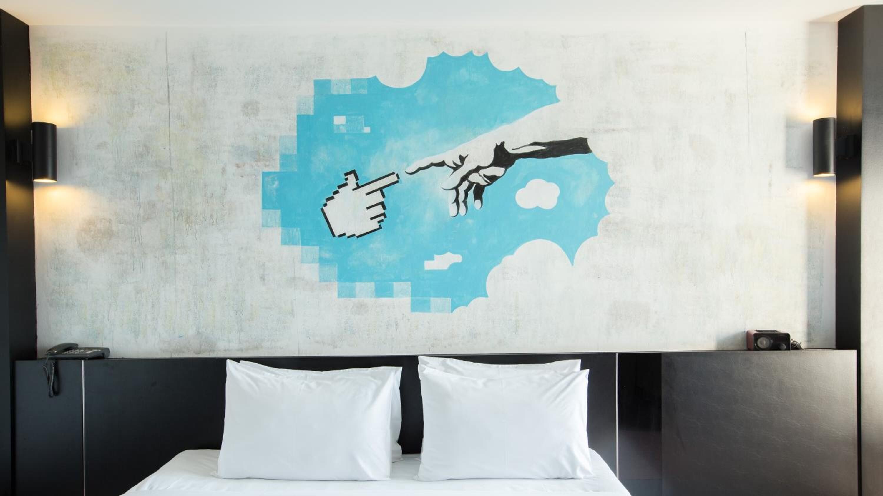 Habitación de hotel con una cama, dos almohadas blancas, un cuadro pixelado en la pared y dos lámparas de pared.