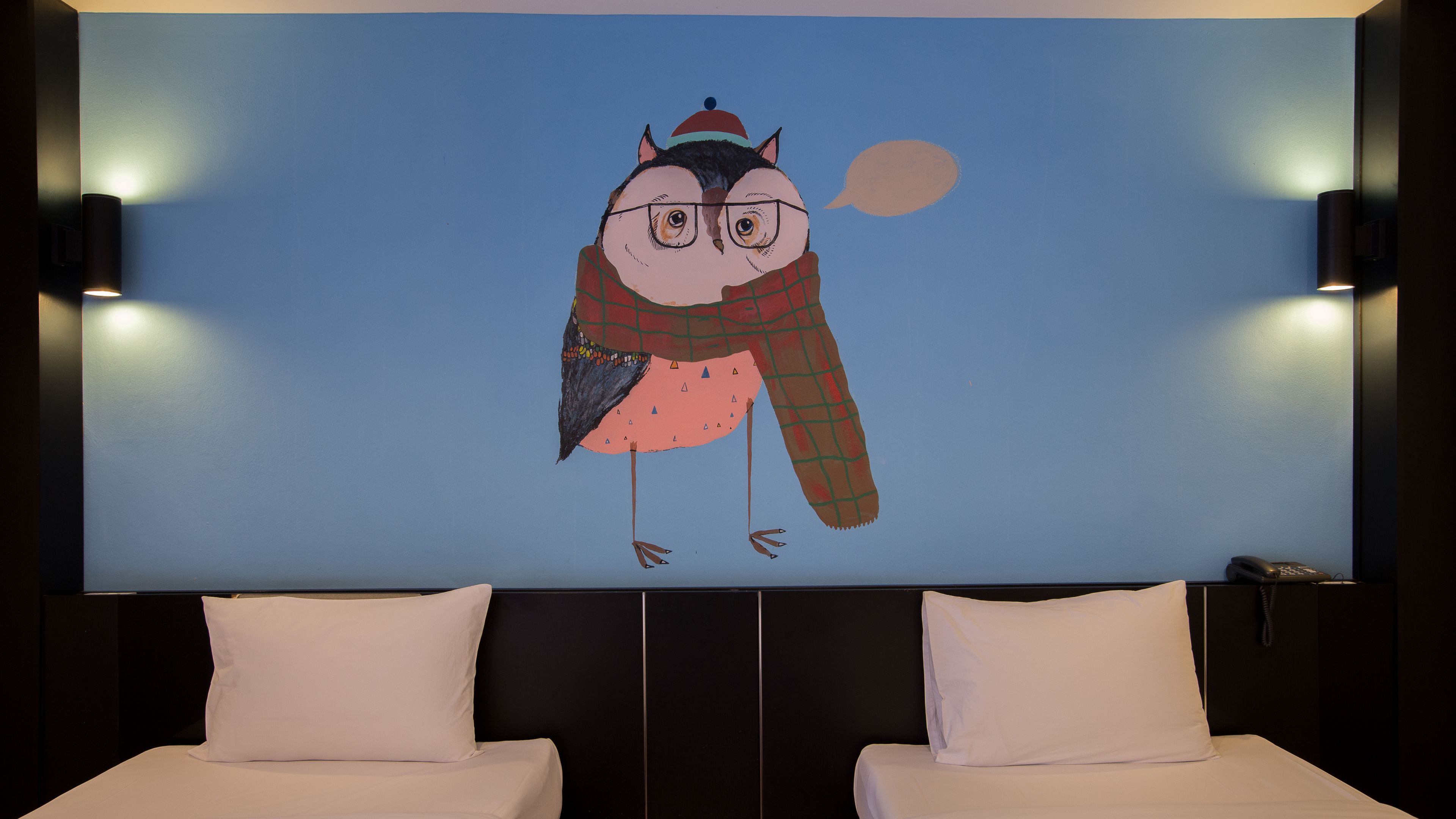 Camera d'albergo con un grande affresco raffigurante un gufo, due letti bianchi e un telefono sul comodino.