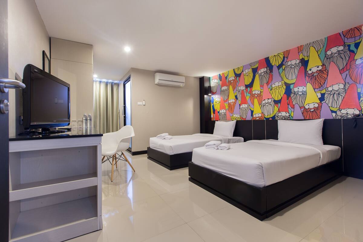Quarto de hotel com televisão, duas camas, uma escrivaninha e um mural colorido na parede.