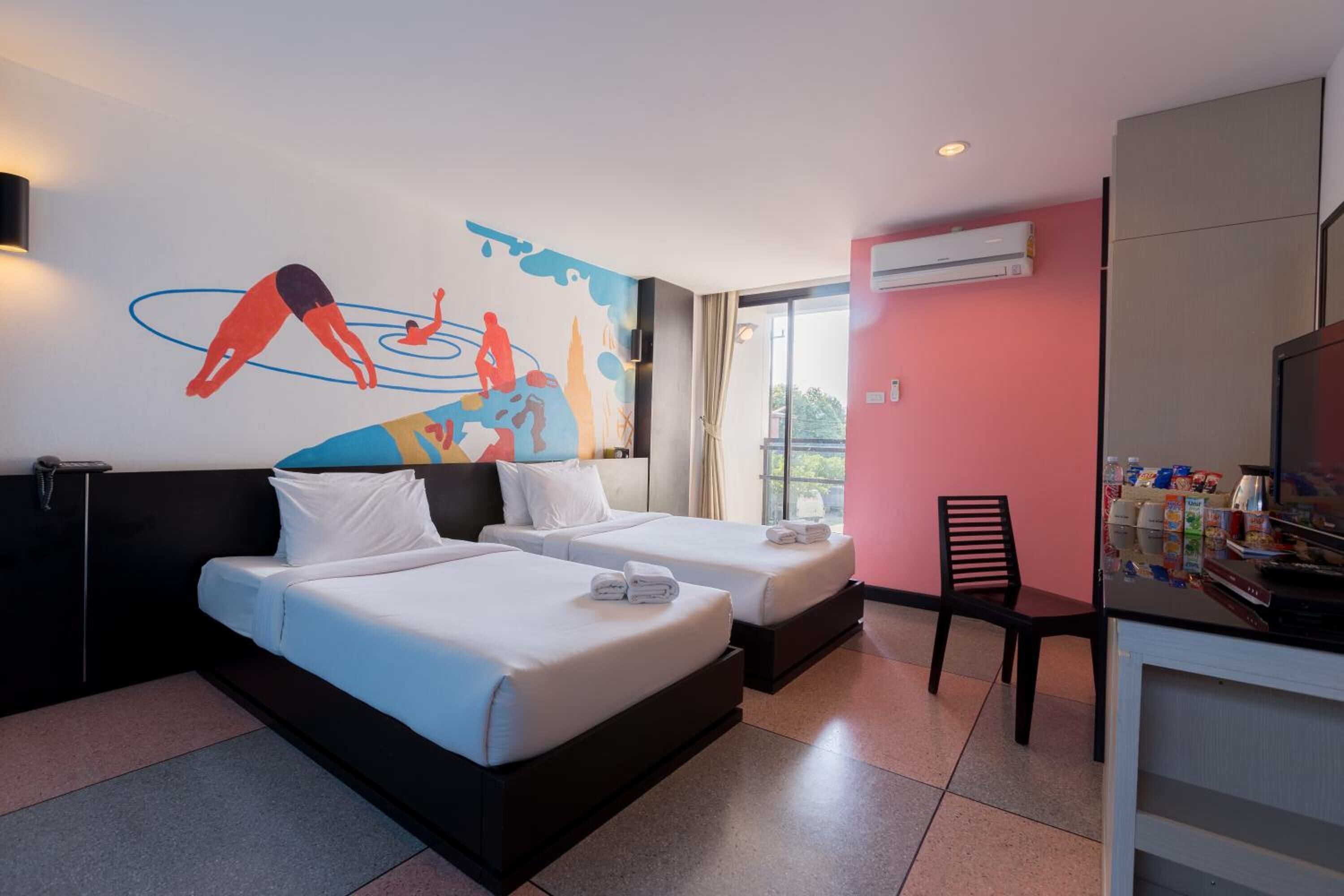 A hotel room with two beds, a desk with a chair, a wall mural, and an air conditioning unit.