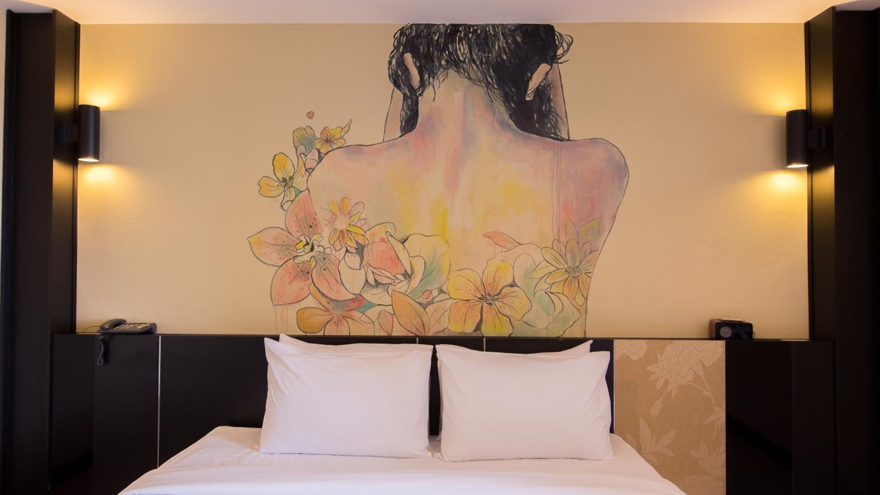 A hotel room with a large bed, two bedside lamps, and a wall mural of a woman with flowers.