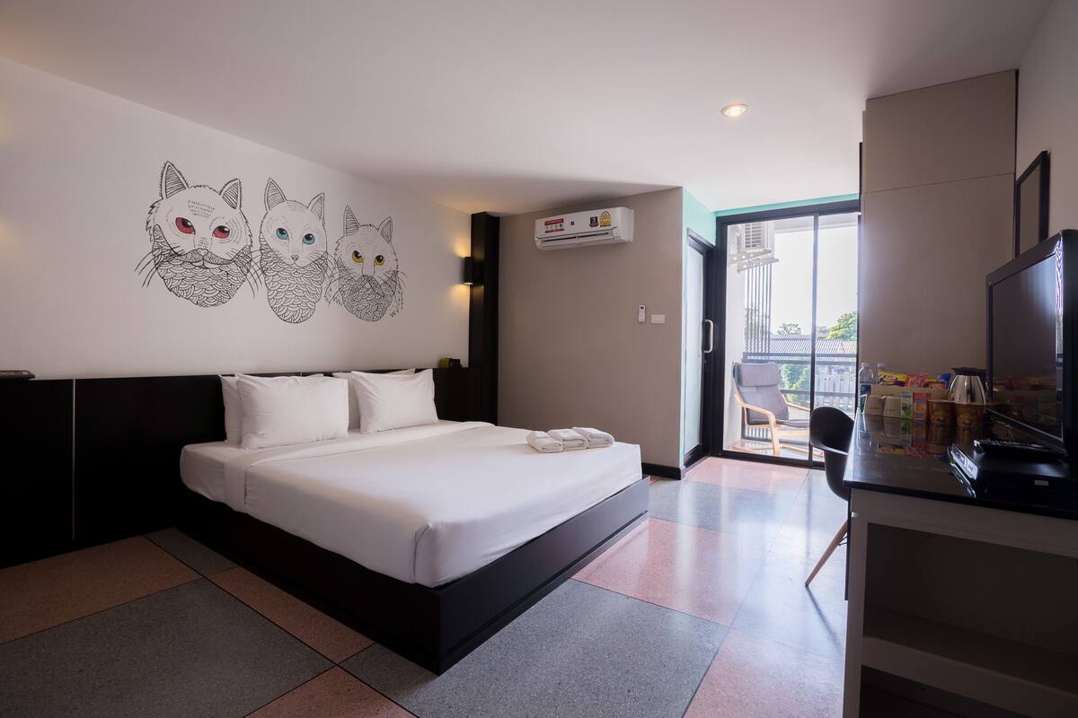 Quarto de hotel com uma cama grande, uma escrivaninha com televisão e uma parede com ilustrações de gatos.