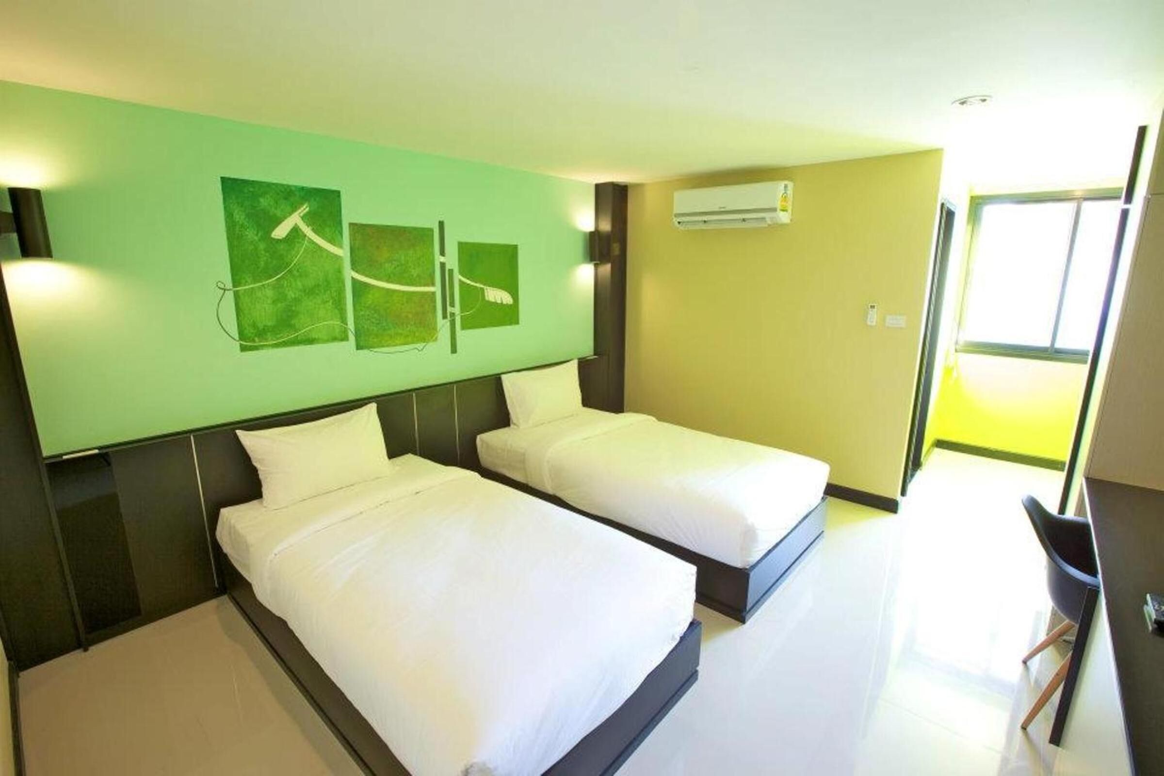 Habitación de hotel con dos camas, una pared verde con obras de arte, un equipo de aire acondicionado y una ventana.