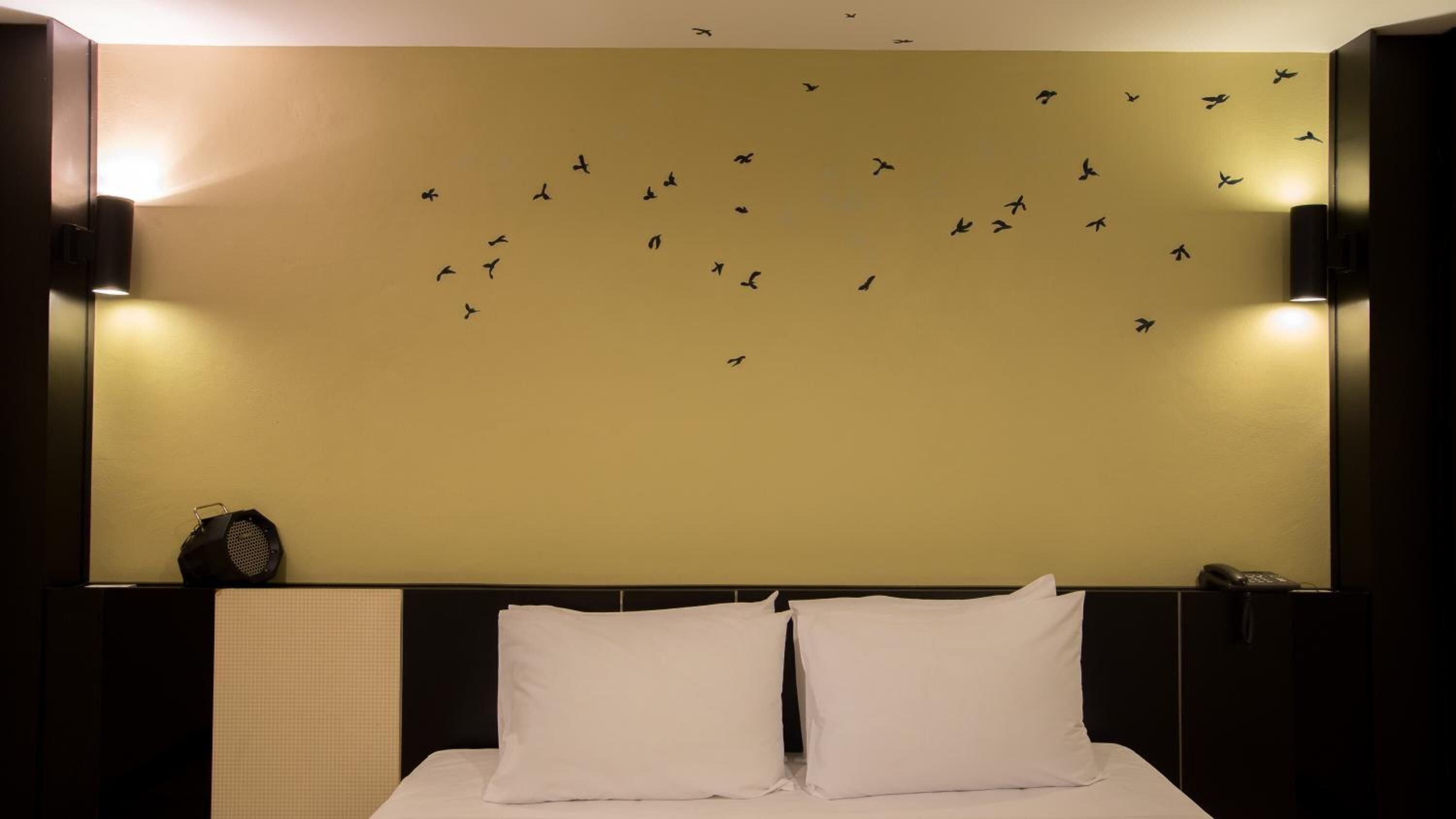 Habitació d'hotel amb un llit gran, almoines blanques, un altaveu fixat a la paret i una calcomania mural de diferents ocells.