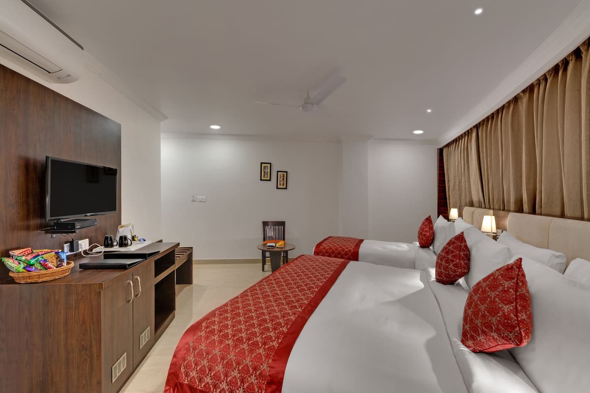 A hotel room with a TV, a desk with snacks, two beds with red pillows, a ceiling fan, and a window with curtains.