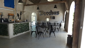 Un vaste espace dédié à la salle à manger, doté d’un haut plafond et de poutres apparentes, comprenant une table centrale entourée de chaises, un comptoir avec des appareils de cuisine et un mur sur lequel est inscrit le nom « Bocchus ».