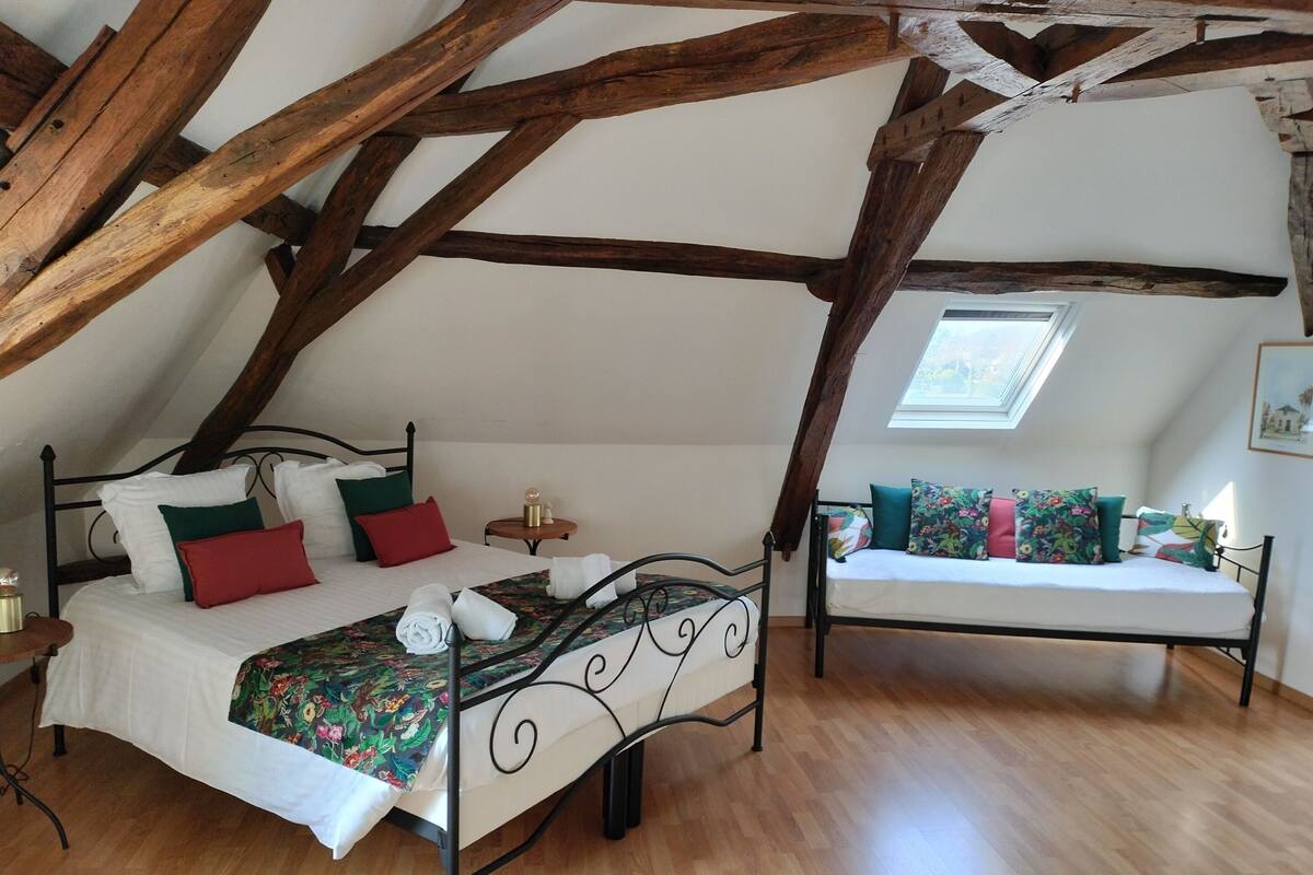 A bedroom with a bed, a daybed, and a small table. The room has wooden beams on the ceiling and a skylight.