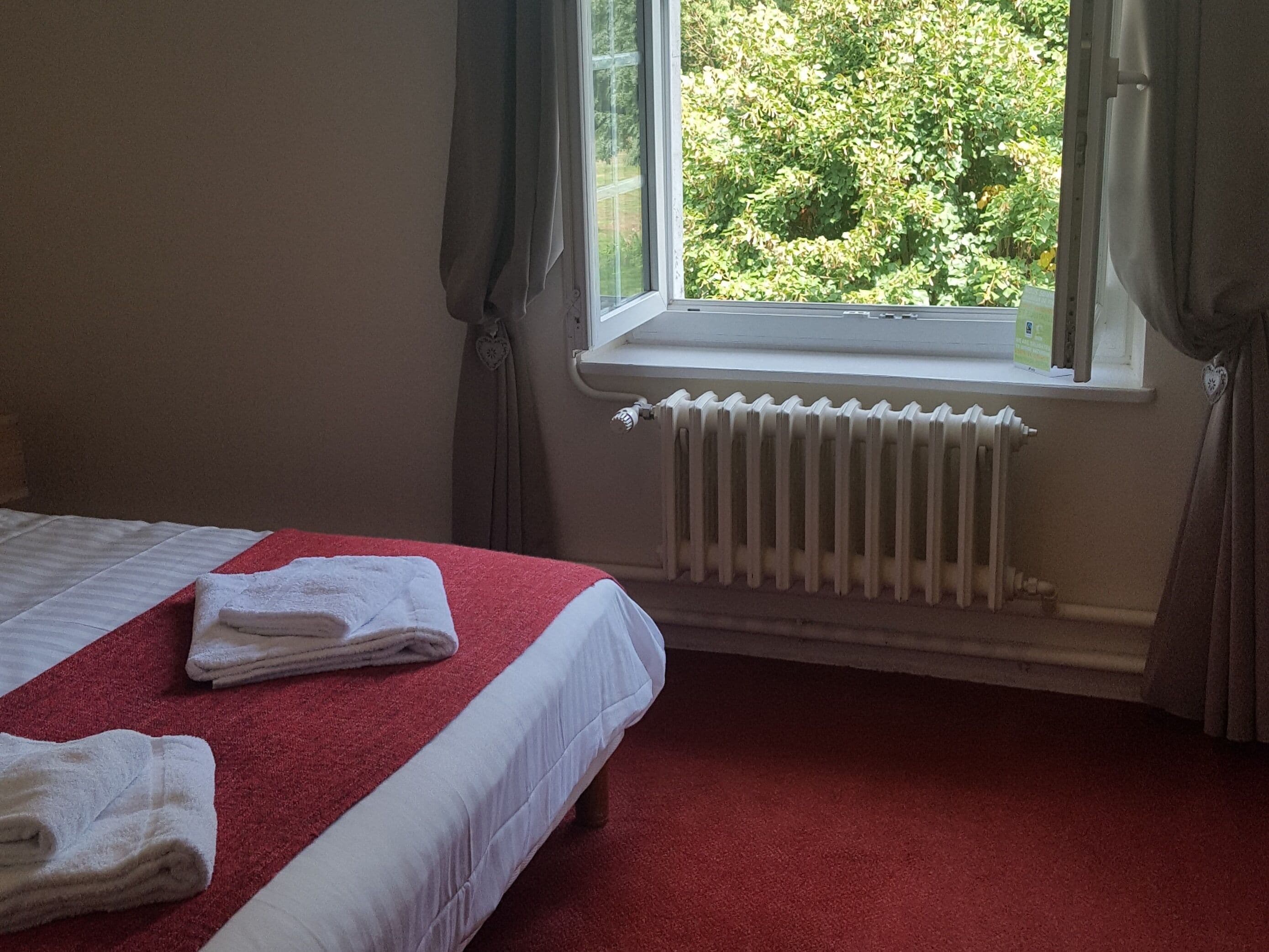 Quarto de hotel com cama, carpete vermelho, roupa de cama branca, janela com cortinas e radiador.