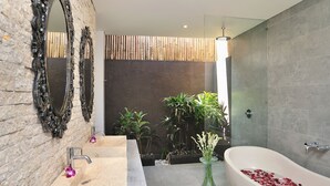 A modern bathroom with a large bathtub, a marble sink, and two mirrors. There are plants and a vase with flowers on the counter. - Villa Flores (Seminyak)