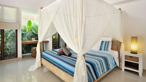 A four-poster canopy bed with a striped blue and white bedspread, a bedside table with a lamp, and a view of greenery through a window. - Villa Flores (Seminyak)