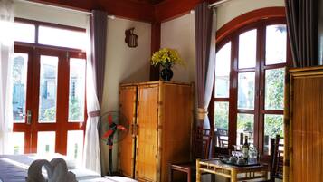 A hotel room with a large bed, a wooden wardrobe, a dining table with chairs, and a fan.