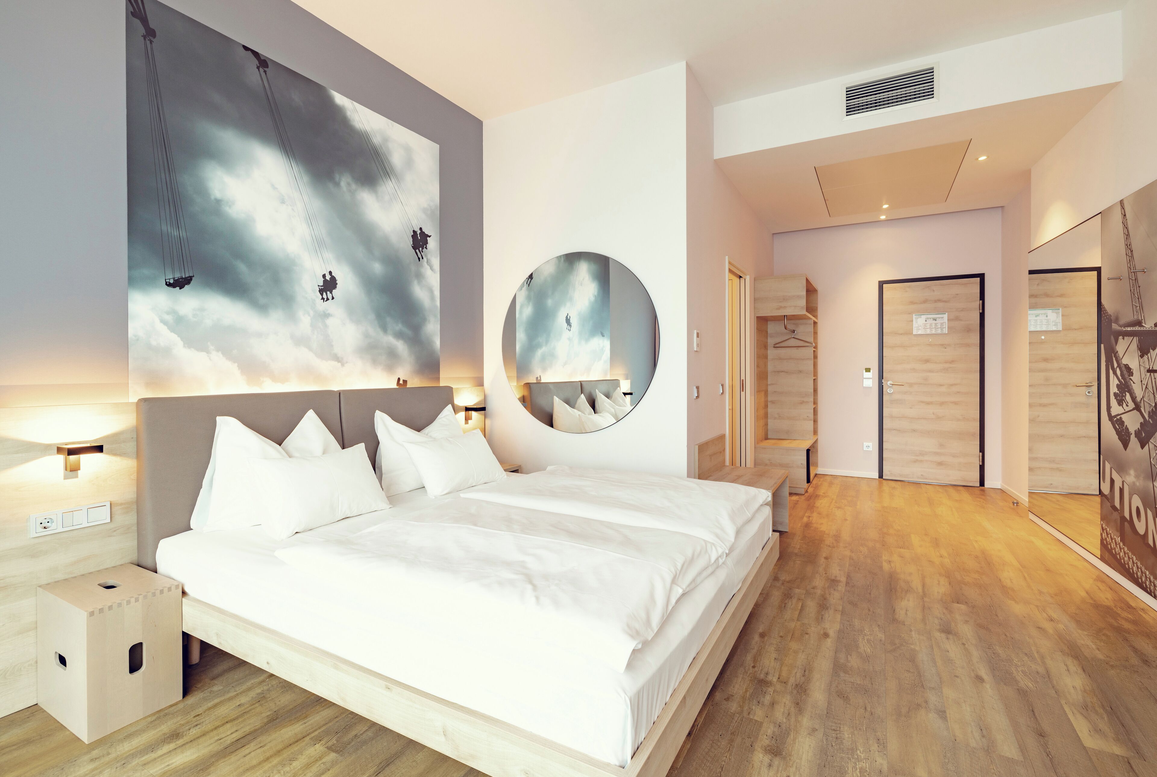 Moderni hotellihuone, jossa on suuri sänky, seinälle kiinnitetty pyöreä peili ja taulu, jossa on kuvattuna taivas pilvineen ja hahmoineen.