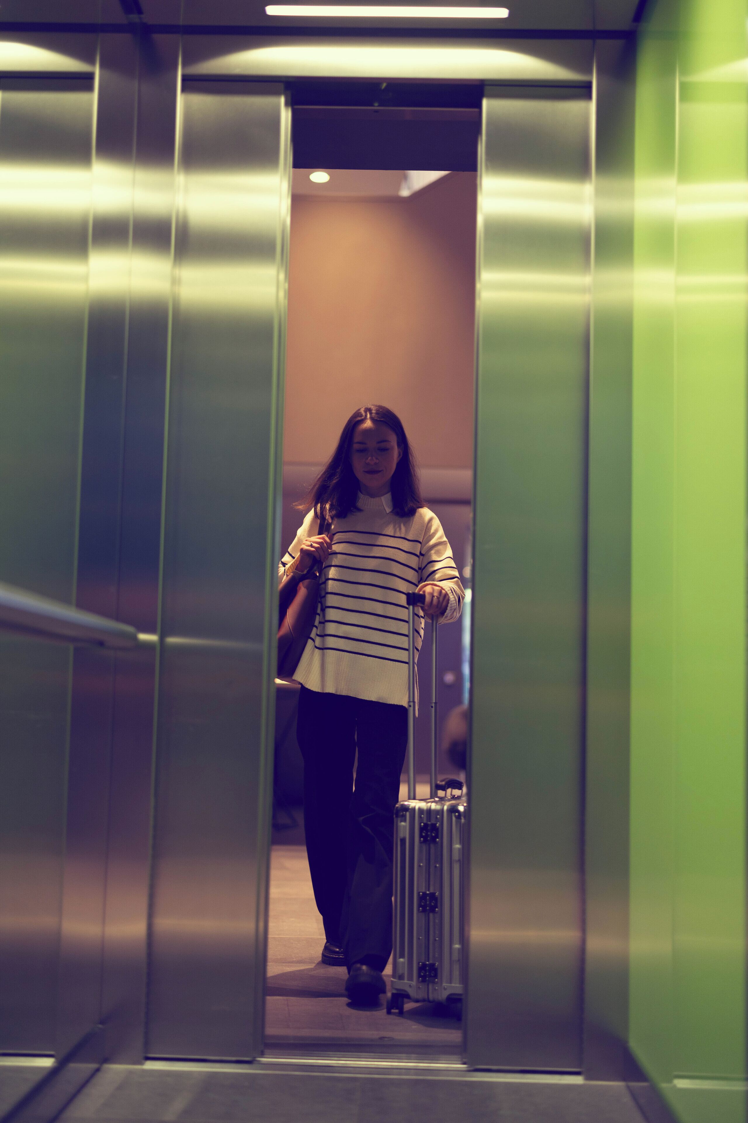 A person in a striped sweater and dark pants walking towards an elevator in a modern building.